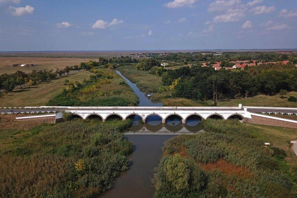 Белый каменный мост с большим количеством арок, проложенный через небольшую речку.